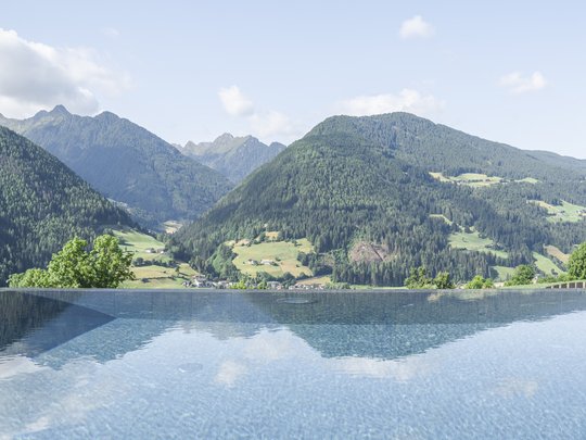 Cercate un hotel a Vipiteno con spa? Benvenuti al Lahnerhof! Piscina a sfioro con vista su montagne verdi e cielo nuvoloso