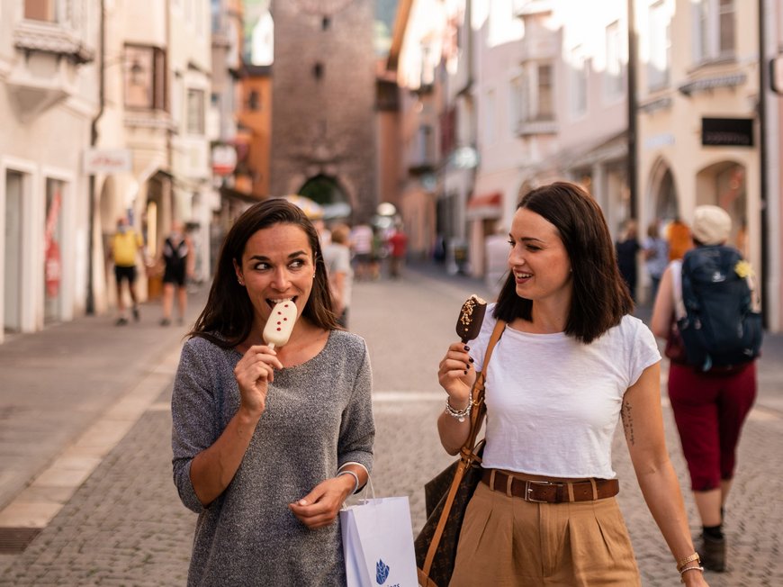 Escursioni sul Monte Cavallo e tanto altro Due donne mangiano gelato e fanno shopping in una città storica con campanile.