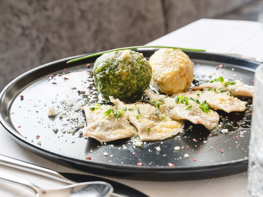 Benvenuti nel ristorante del nostro hotel con spa a Vipiteno Piatto nero con ravioli, due canederli e erbe su tovaglia bianca
