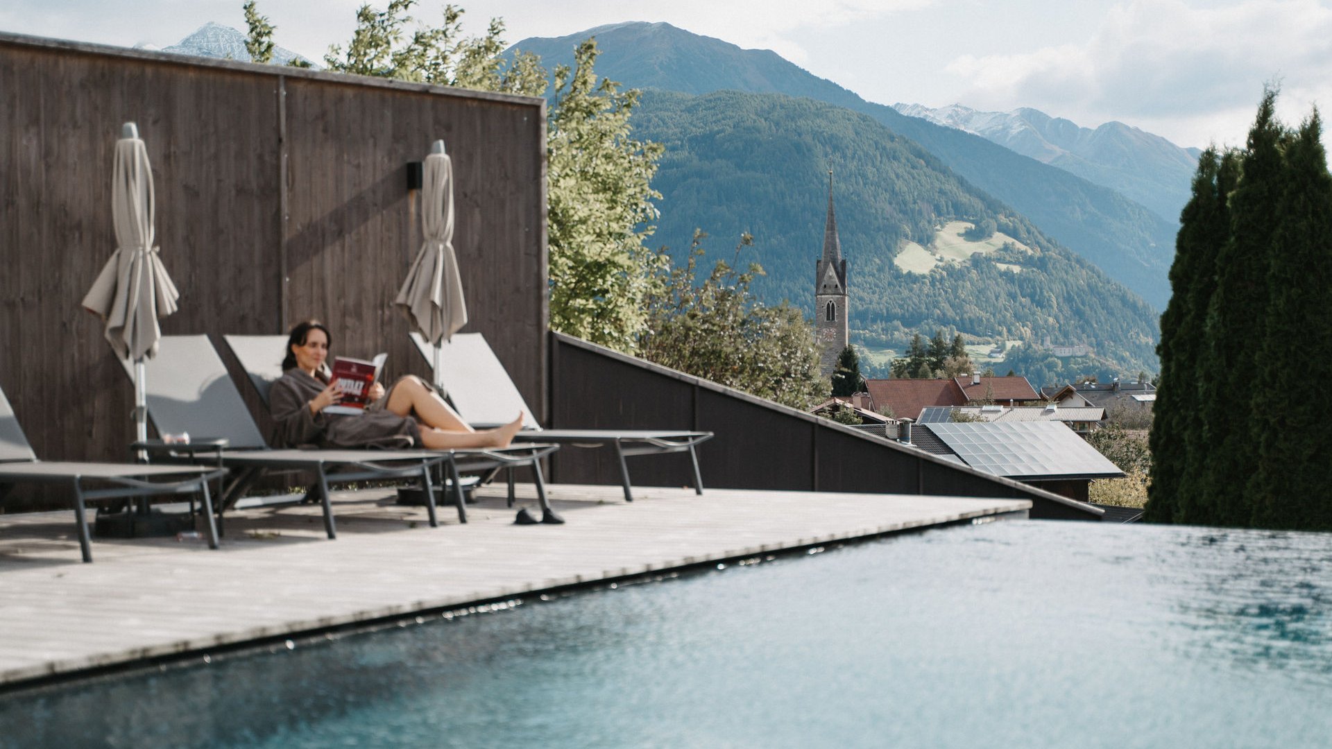 Lahnerhof: Natur- und Wellnesshotel bei Sterzing Frau entspannt auf Liegestuhl am Pool mit Bergblick und Kirchturm im Hintergrund