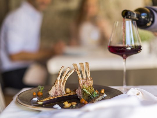 Benvenuti nel ristorante del nostro hotel con spa a Vipiteno Costolette di agnello su un piatto con un bicchiere di vino rosso sullo sfondo