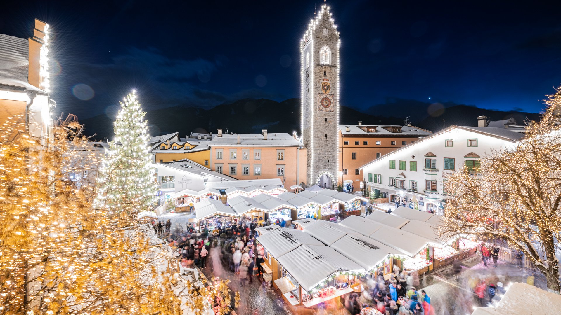 Lahnerhof: Natur- und Wellnesshotel bei Sterzing Belebter Weihnachtsmarkt bei Nacht mit beleuchteten Ständen, geschmückten Bäumen und Uhrturm