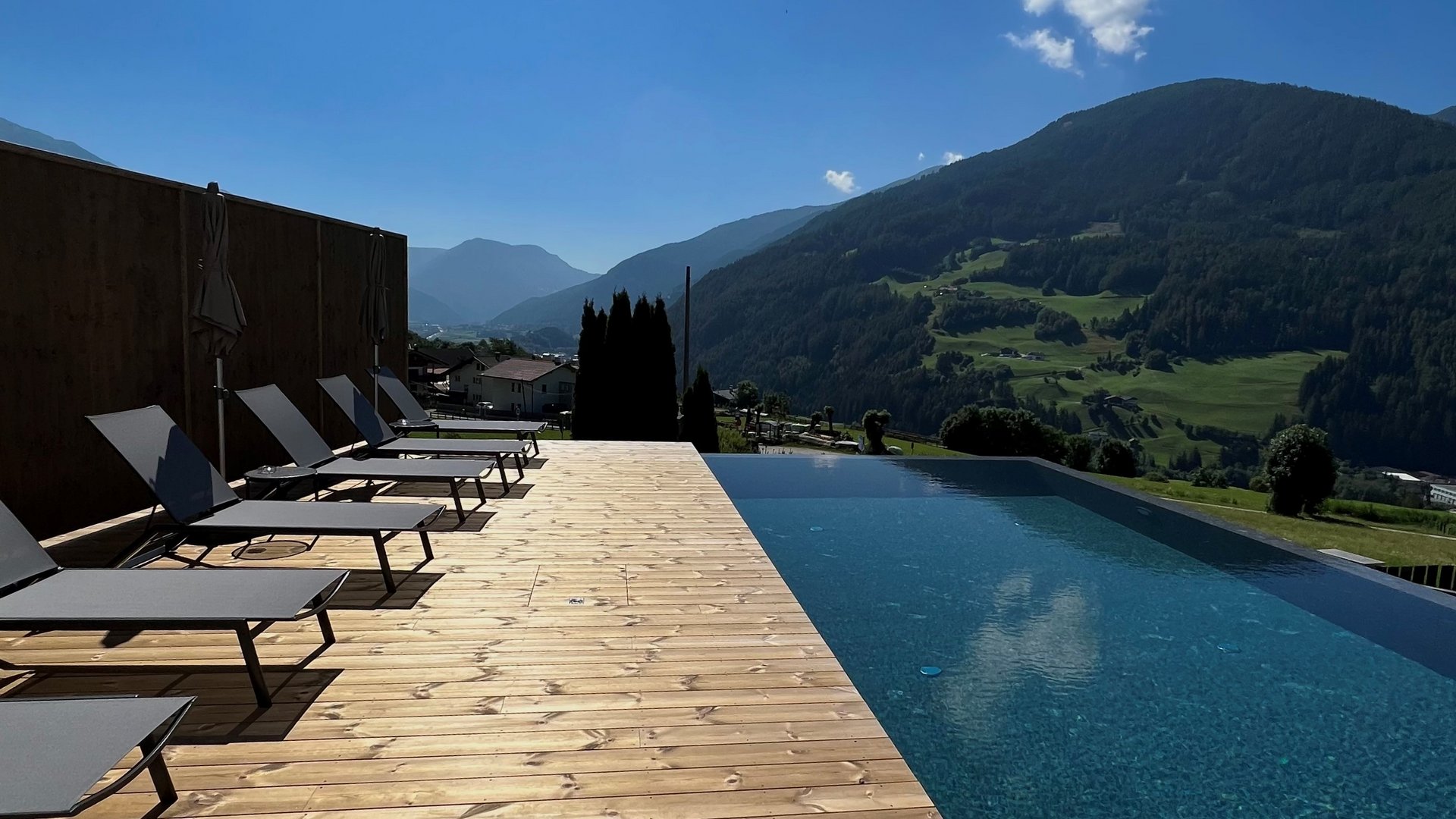 Cercate un hotel a Vipiteno con spa? Benvenuti al Lahnerhof! Piscina a sfioro con vista sulle montagne e lettini su terrazza in legno sotto il sole