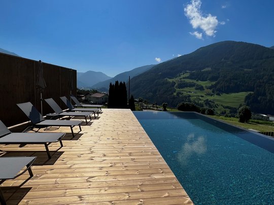 Cercate un hotel a Vipiteno con spa? Benvenuti al Lahnerhof! Piscina a sfioro con vista sulle montagne e lettini su terrazza in legno sotto il sole